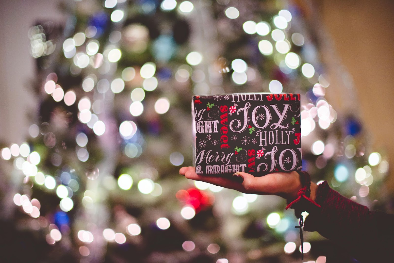 An outstretched hand presenting a wrapped holiday gift with a christmas tree in the background