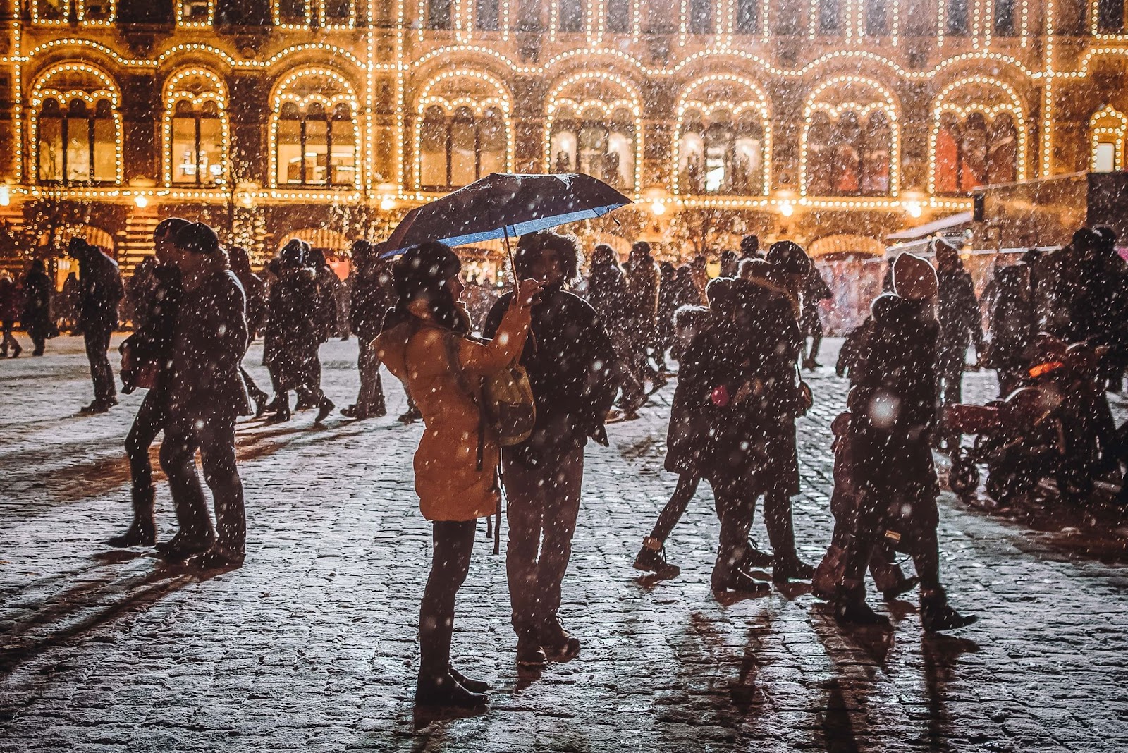 People walking on a busy street in the snow with christmas lights adorned the building in the background