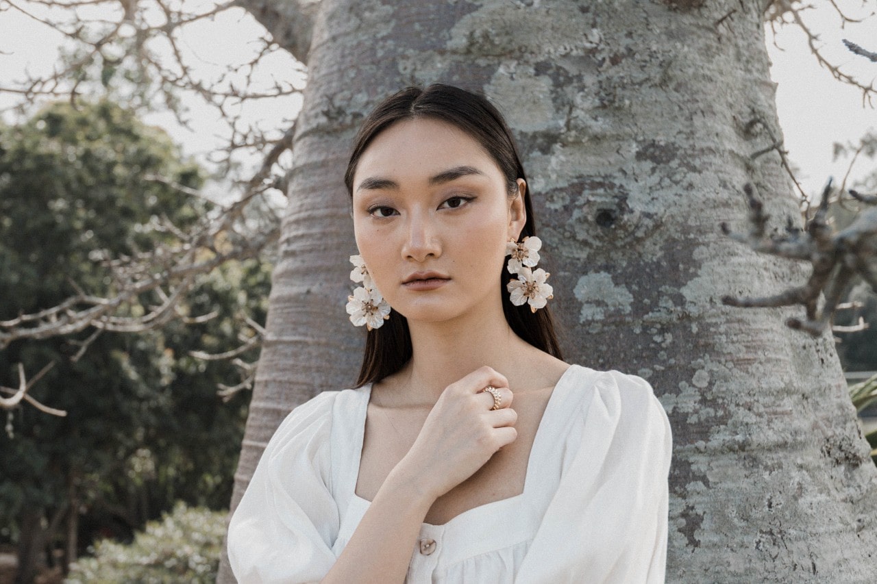 A woman standing in front of a tree wearing a pair of large floral earrings.