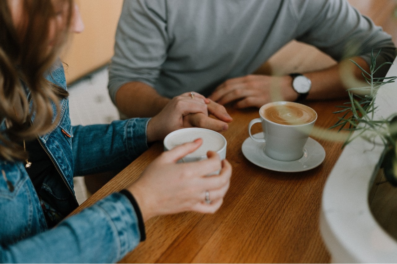 An engaged couple sips coffee at a restaurant while holding hands.