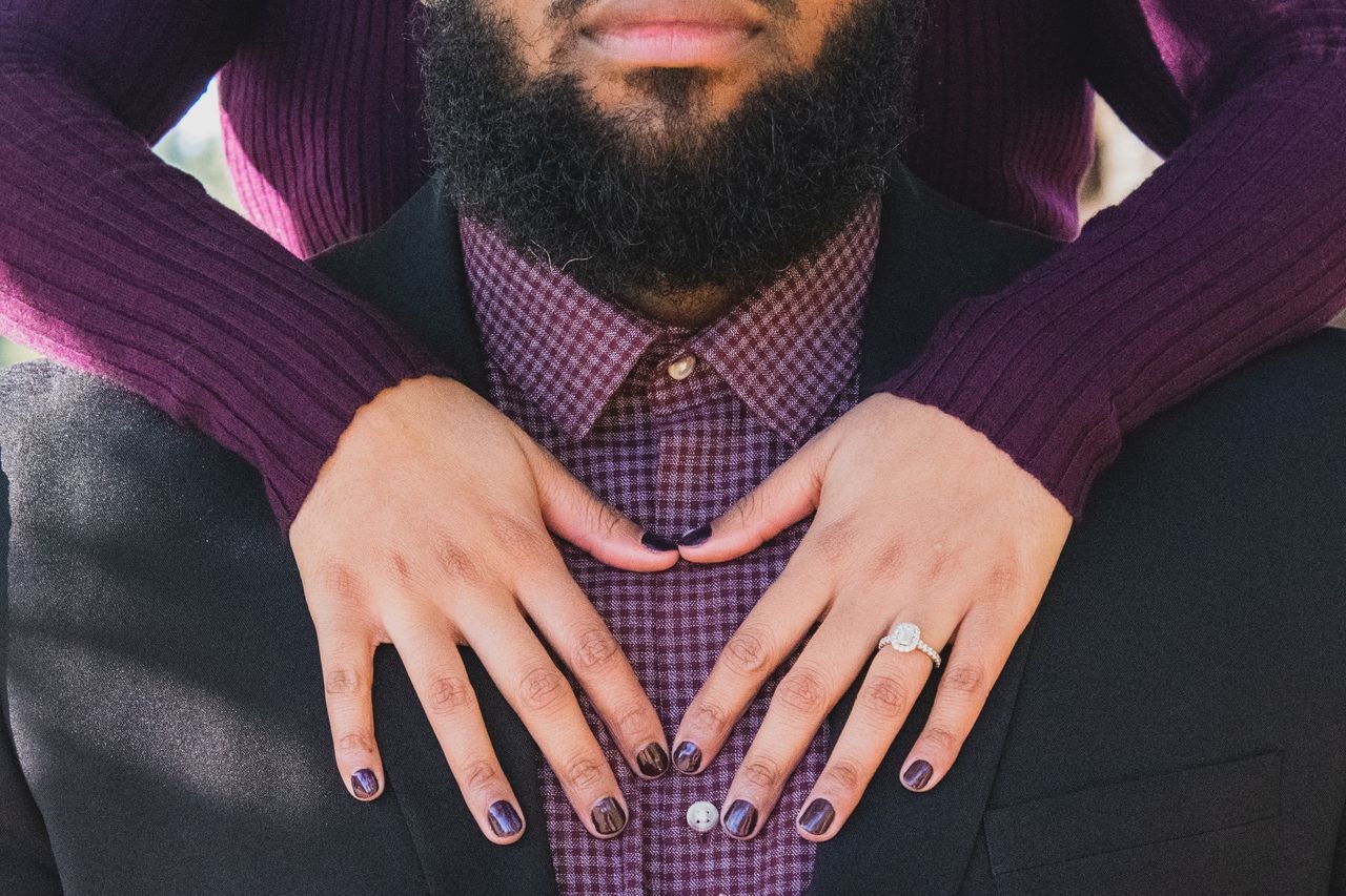 A woman hugs her fiance from behind while showing off her oval halo engagement ring.