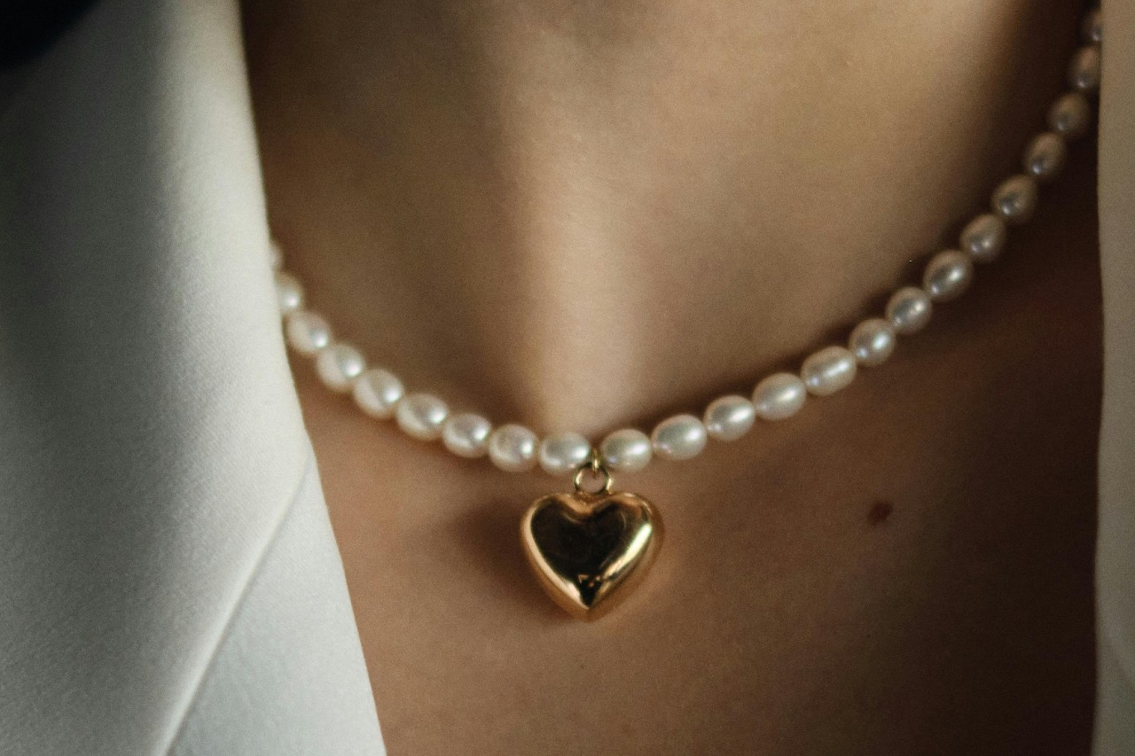 A close-up of a woman’s neckline, adorned with a pearl necklace with a heart pendant.