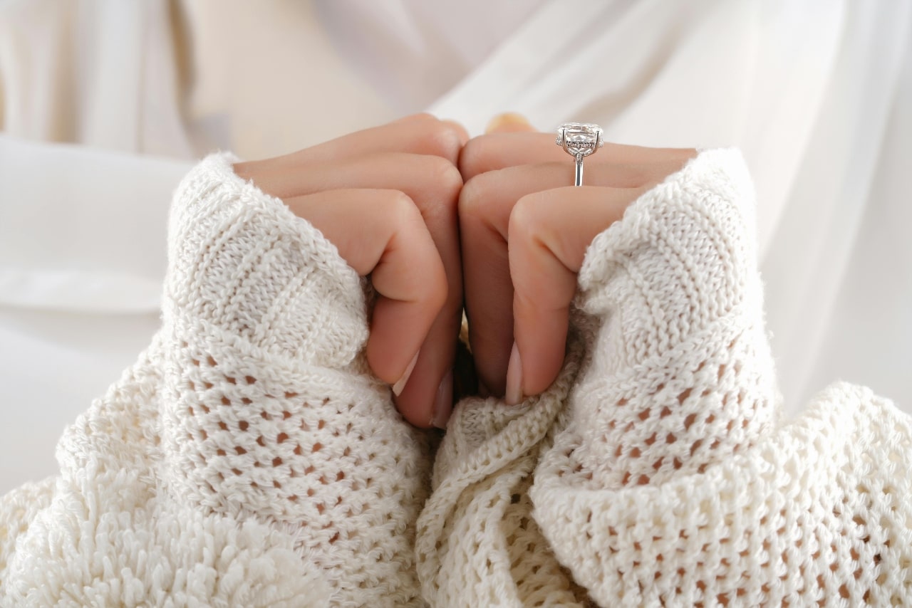 A close-up of a woman wearing a comfy sweater, an elegant engagement ring on her finger.