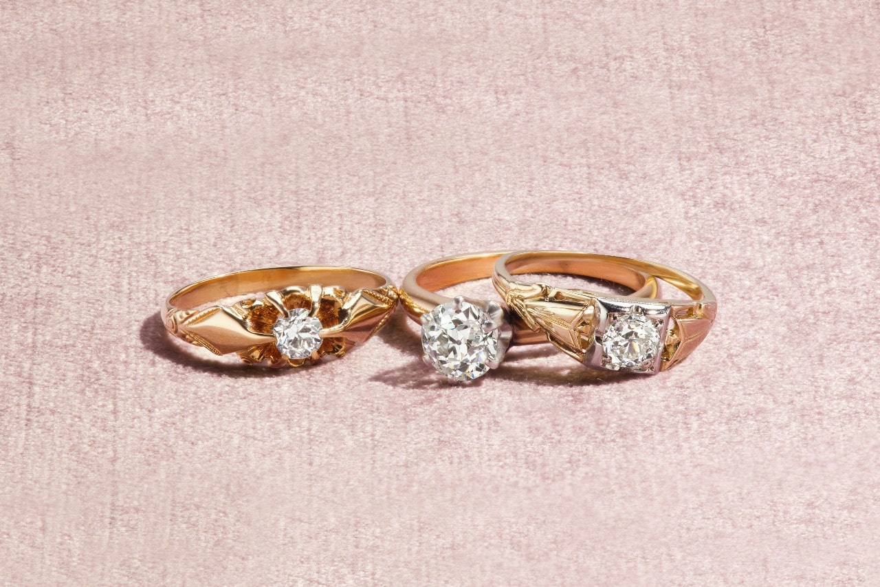 A close-up of three elegant solitaire engagement rings displayed on a plush pink backdrop.